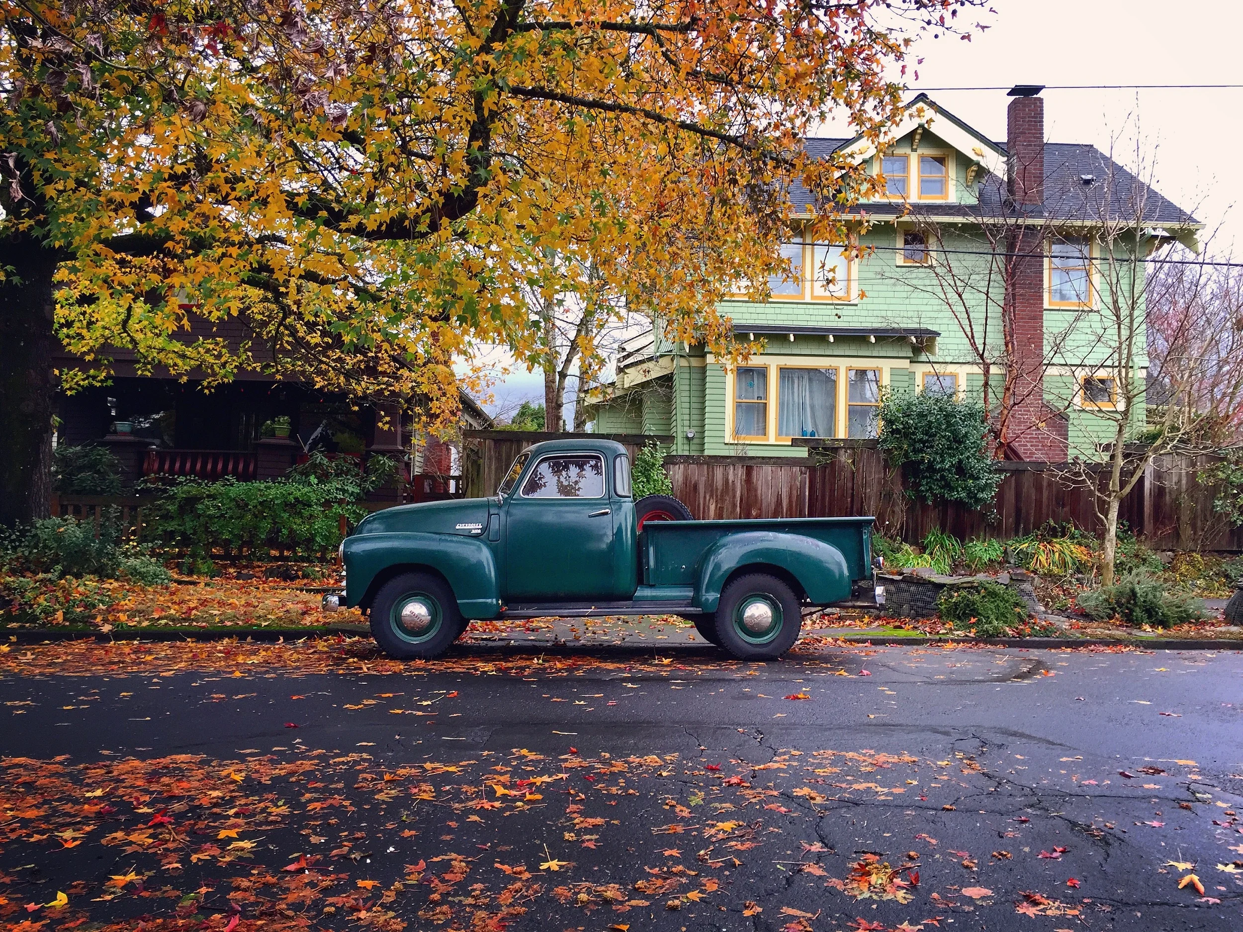 A teal vintage truck parked beside a green house in autumn